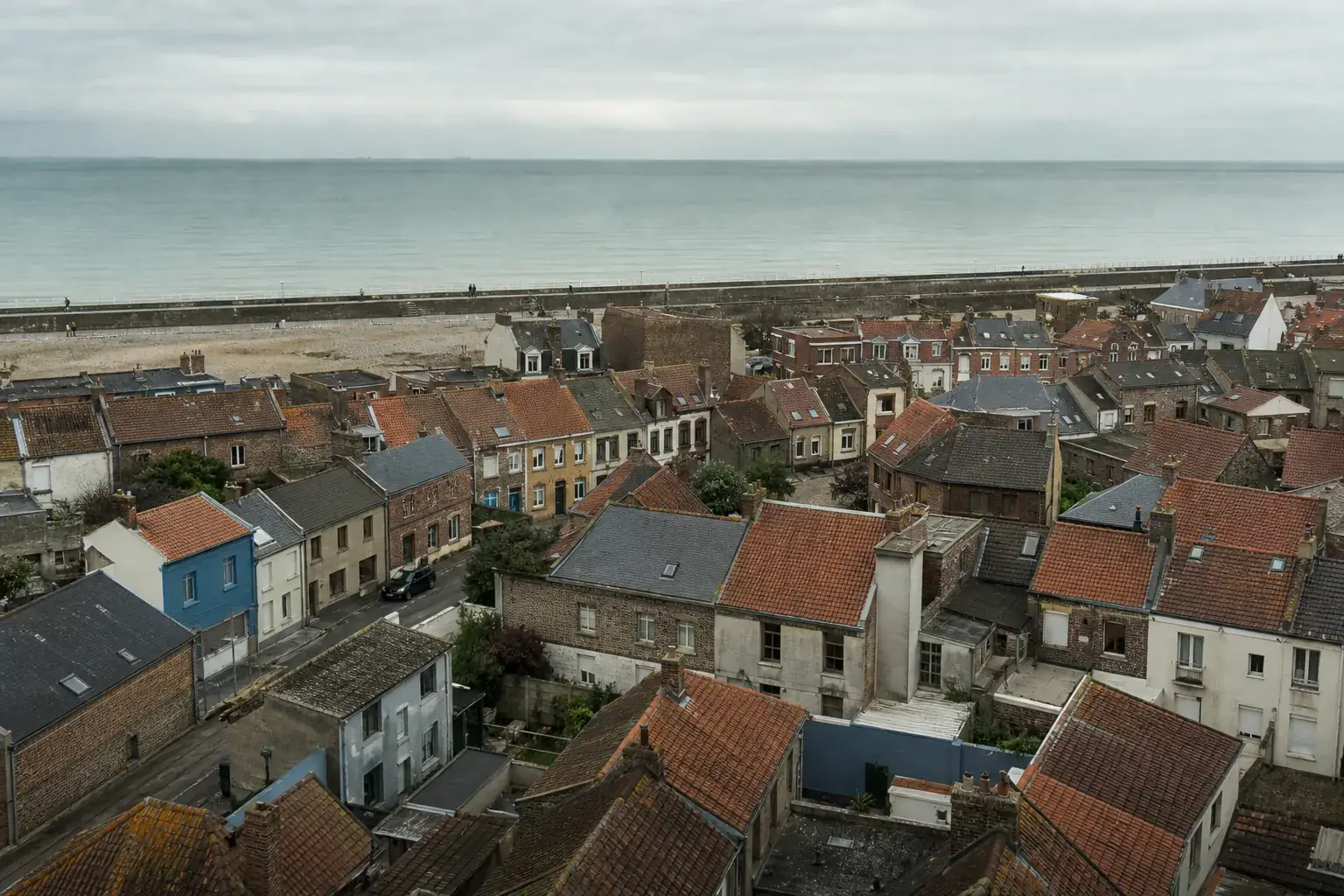Vue aérienne basse de la côte d’Opale près de Calais, toitures de brique et d’ardoise, digue et mer du Nord au fond, ciel couvert, photographie documentaire