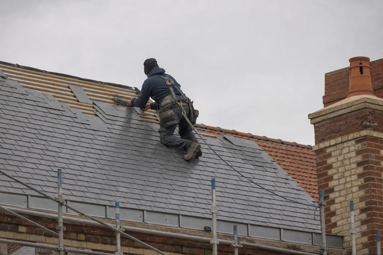 Couvreur en hauteur posant des ardoises sur un versant de toiture, harnais de sécurité, échafaudage, ciel gris