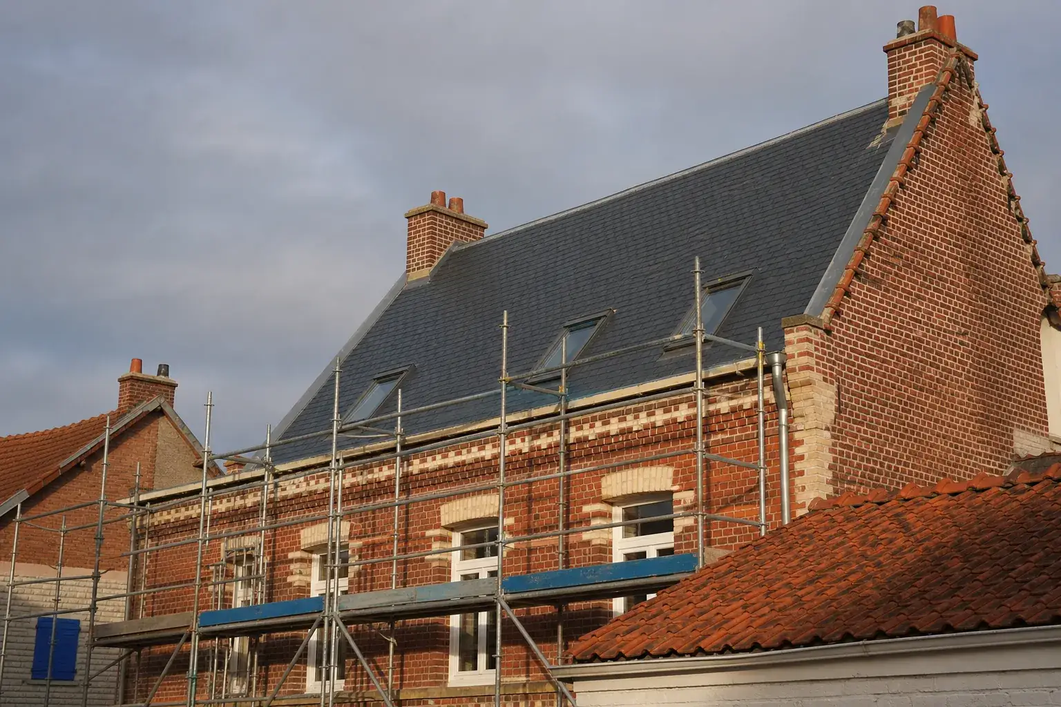 Toiture en ardoise fraîchement refaite sur une maison de brique du nord de la France, échafaudage encore en place, ciel gris de Manche, lumière rasante de fin de journée, photographie documentaire sans personne