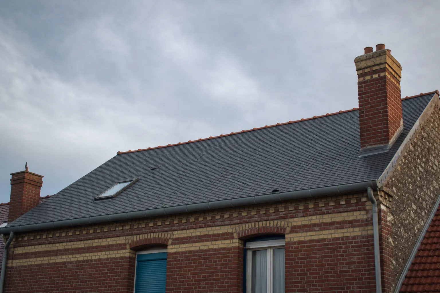 Toiture en ardoise d’une maison de brique du nord de la France vue en contre-plongée depuis la rue, ciel bas et gris de Manche, lumière rasante de fin de journée, aucune personne, photographie documentaire